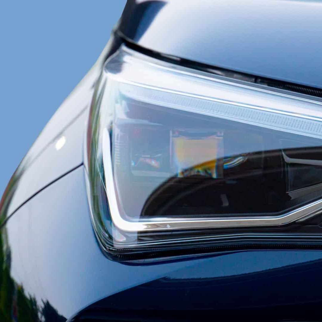 Close-up of a car headlight assembly against a light blue background. Headlight is clear with internal details visible and the car dark blue paint is reflected on its surface.